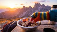 Person wearing colorful striped gloves reaching for snacks in bowl at mountain campsite during golden hour sunset with alpine peaks in background.