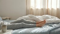 Woman in gray leggings and white shirt relaxing on bed with white bedding near bright window with sheer curtains, morning leisure and comfort concept.