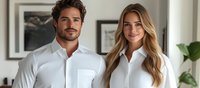Young Caucasian couple wearing matching white button-up shirts smile at camera in modern bright interior setting with framed artwork on wall. Professional lifestyle portrait.
