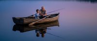 Father and son fishing together from wooden boat on calm lake at sunset with perfect water reflections creating peaceful family bonding moment outdoors.