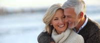 Affectionate mature couple embracing outdoors, wearing cozy sweaters. Blonde woman and silver-haired man sharing intimate moment against blurred coastal background.