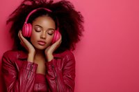 Young African American woman with natural curly hair wearing red leather jacket and pink headphones enjoying music with eyes closed against pink background.