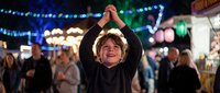 Young boy with arms raised celebrating at colorful nighttime carnival with festive lights and crowd enjoying fair atmosphere.