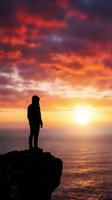 Silhouetted figure standing on rocky cliff overlooking ocean at dramatic sunset with vibrant orange and red sky, conveying contemplation, adventure, and inspirational themes for motivational content.