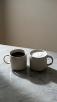 Two white ceramic mugs on marble surface, one filled with black coffee, the other with white milk or cream, casting shadows in natural light for beverage and lifestyle content.