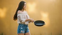 Young Hispanic woman in white embroidered top and denim shorts holding cast iron skillet outdoors during golden hour sunset for cooking and lifestyle content.