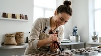 Young woman with brown hair in a bun, Caucasian ethnicity, wearing glasses and beige apron, working on jewelry or craft project at table in bright studio space.