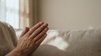 Elderly person's hands clasped together in prayer near a sunlit window, expressing faith, hope, and spirituality in a calm home setting.