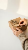 Person with light skin tone holding kraft paper wrapped gift box with eucalyptus decoration and twine, wearing beige knit sweater. Suitable for holiday, gift-giving, sustainable packaging, and celebration content.