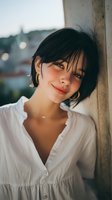 Young woman with short dark hair and gold hoop earrings leaning against wall in white button-up blouse, soft sunset lighting creating warm atmosphere for lifestyle portraits.