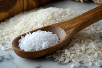 Raw white rice grains in wooden spoon on marble surface, scattered uncooked rice in background, macro food photography for cooking and cuisine concepts.