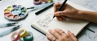 Artist hand sketching creative designs in notebook with watercolor paints and brushes on marble desk for artistic projects and illustration work.