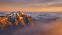 Snow-capped mountain peak rising above a sea of clouds at golden sunset, dramatic alpine landscape with warm orange and pink sky tones.