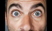 Close-up portrait of a Caucasian man with wide-open blue eyes expressing extreme surprise or shock against dark background.