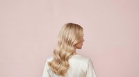 Young woman with long blonde wavy hair, fair complexion, wearing white top, shown in profile against soft pink background for beauty and hair care marketing.