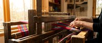 Senior craftsman weaving colorful plaid fabric on traditional wooden loom in sunlit workshop. Hands working with red and blue threads in artisan textile studio.
