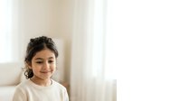 Young woman with dark hair and olive complexion wearing a cream knit sweater, smiling at camera in bright, minimalist interior space with natural window light for portrait and lifestyle use.