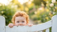 Adorable toddler with curly blonde hair peeking over white wooden fence in sunny garden setting with soft bokeh background for family lifestyle content.