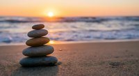 Balanced stone stack on sandy beach at golden sunset with ocean waves creating peaceful zen meditation atmosphere for wellness and mindfulness practices.