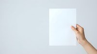 Female hand holding blank white paper sheet against neutral gray background, suitable for mockups, presentations, and advertising campaigns.