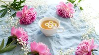 Herbal chamomile tea in elegant cup surrounded by pink peonies and white flowers on marble surface for relaxation and wellness lifestyle concepts.