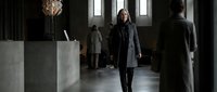 Professional woman in dark coat walking through modern office lobby with colleagues in background during business day.
