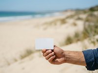 Cropped view of male hand holding blank white card at sandy beach with ocean and dunes in background, ready for custom text or branding on vacation or travel marketing materials.