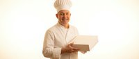 Middle-aged male chef with light skin tone wearing white chef's uniform and tall chef's hat, holding an open cardboard box against neutral background.