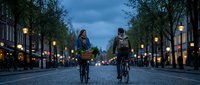 Young couple cycling together on cobblestone street at dusk with warm street lights creating romantic urban atmosphere for evening date adventure.