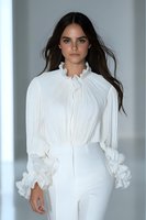Young Caucasian woman with long dark hair wearing elegant white ruffled blouse with Victorian-style collar and white pants against light background. Fashion portrait.