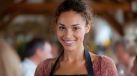 Young mixed race female restaurant worker wearing black apron and pink sweater smiling warmly at camera in busy dining room setting with natural lighting.