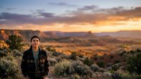 Young woman with brown hair and fair complexion wearing a dark patterned jacket stands in desert landscape at sunset, gazing toward golden sky with distant mesas and warm light.
