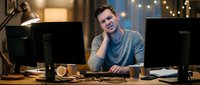 Young Caucasian male programmer working late at night in modern office with dual monitors, coffee cup and warm lighting creating productive workspace atmosphere.