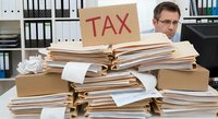 Overwhelmed businessman with glasses behind massive pile of tax documents and paperwork in organized office environment during tax season preparation.