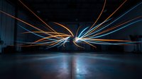 Streaks of orange and blue light converge in empty warehouse space, creating dynamic motion effect for technology and innovation concepts.