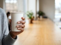 Young Black woman holding blank white paper cup in modern office environment with wooden table and blurred background, suitable for beverage branding and corporate lifestyle content.