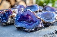 Blue and purple agate crystal slices with natural banded patterns and rough edges displayed on dark stone surface, showing detailed mineral formations.