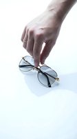 Hand picking up stylish aviator sunglasses with gold metal frame and blue tinted lenses on a white background with soft shadow.