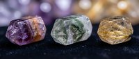 Three raw gemstone crystals in purple amethyst, green prehnite and golden rutilated quartz arranged on dark surface with bokeh background.