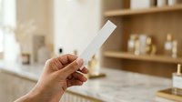 Cropped hand holding blank white product box in modern kitchen interior with wooden shelves and natural lighting for packaging mockup.
