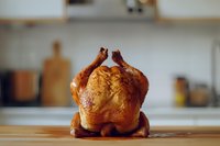 Whole roasted chicken with golden brown crispy skin sitting on wooden kitchen counter with blurred culinary background, ready for cooking content and food styling.