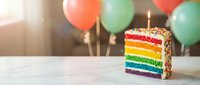Colorful rainbow layer cake slice with lit candle on white table, festive balloons in background for birthday celebration party dessert.