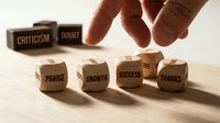 Hand placing wooden cube with positive words while negative criticism and doubt blocks remain in background on wooden surface.