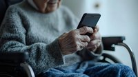 Senior Caucasian person in gray sweater using smartphone while sitting in chair, close-up view of hands holding mobile device. Represents modern technology adoption.