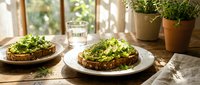 Fresh avocado toast with microgreens on rustic wooden table in bright kitchen setting with natural sunlight and potted herbs for healthy breakfast.