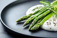Fresh green asparagus spears on black plate with creamy white sauce and cracked pepper, garnished with herbs. Elegant food styling for restaurant menu.