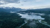 Aerial view of misty mountain valley with winding river through dense forest, snow-capped peaks and low clouds creating dramatic wilderness landscape.