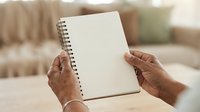Mature woman with dark skin tone holding open blank spiral notebook with white pages indoors, suitable for education, planning, journaling, and creative writing projects.