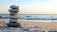 Balanced stack of smooth zen stones on sandy beach at sunset with ocean waves and horizon in soft focus background creating peaceful atmosphere.