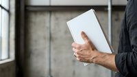 Businessman holding blank white tablet device in modern office space with industrial concrete walls and natural window light.